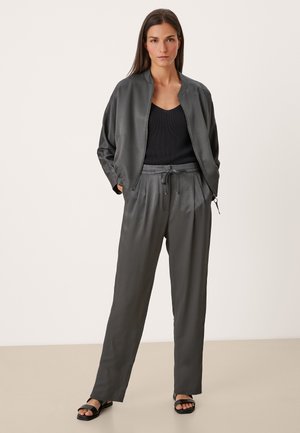 Femme portant une veste bomber en satin gris foncé, un top en tricot noir, un pantalon taille haute assorti et des sandales noires, se tenant avec les mains dans les poches.