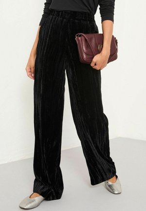 Woman wearing loose black velvet pants, silver slip-on shoes, holding a small dark red leather handbag against a plain background.