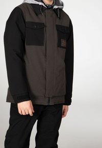Giacca isolata nera e grigia con due tasche sul petto, cappuccio e polsini elastici. Presenta una toppa con logo e una chiusura zip frontale.