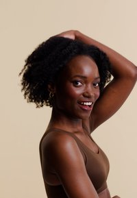 Soutien-gorge de sport marron porté par une personne avec des cheveux noirs courts et bouclés. L'arrière-plan est d'un beige neutre, mettant en valeur le teint de la peau et la texture du tissu.