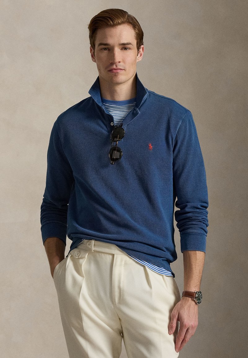 Men's navy blue long-sleeve polo shirt with a collar, featuring a red logo, worn over a striped shirt, paired with cream pants and sunglasses.