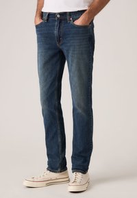 Σκούρο μπλε τζιν με ίσια γραμμή, ξεθωριασμένη υφή, πέντε τσέπες και κούμπωμα με κουμπί, συνδυασμένο με λευκά πάνινα αθλητικά παπούτσια.