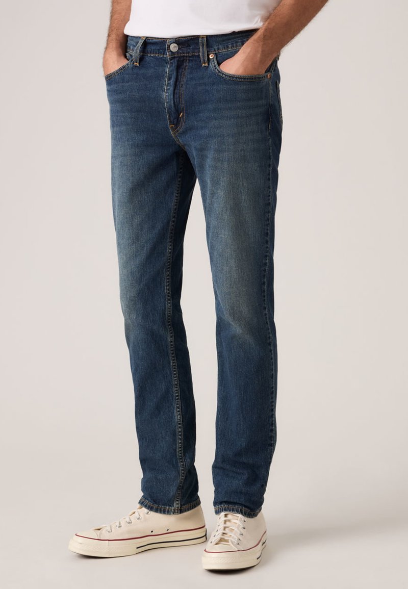 Σκούρο μπλε τζιν με ίσια γραμμή, ξεθωριασμένη υφή, πέντε τσέπες και κούμπωμα με κουμπί, συνδυασμένο με λευκά πάνινα αθλητικά παπούτσια.