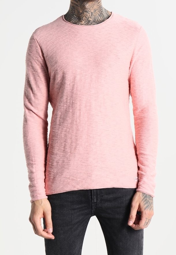 Homme portant une chemise à manches longues rose clair et un jean foncé, avec des tatouages visibles sur le cou et les mains, debout devant un fond uni.