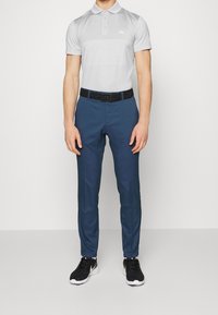 Man som bär en ljusgrå kortärmad pikétröja, mörkblå slim-fit byxor, svart bälte och svarta sportskor, stående mot en neutral bakgrund.