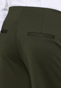 Pantalon sur mesure vert olive avec poches passepoilées à l'arrière, porté avec une chemise blanche rentrée.