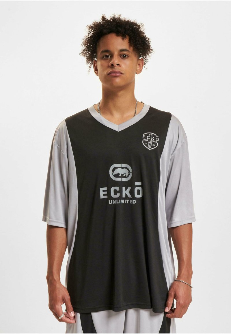 Maillot noir et gris à manches courtes avec un col en V, doté d'un logo sur la poitrine et d'une manche gauche grise contrastante. Fabriqué en tissu lisse.