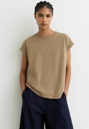 Kvinde med fletninger iført en beige ærmeløs top og marineblå bukser med nålestriber, stående op ad en hvid væg med hænderne i lommerne.