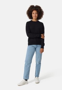 Schwarzer Pullover mit langen Ärmeln und Rundhalsausschnitt, kombiniert mit hellblauen Jeans und weißen Sneakers. Das Modell zeigt eine entspannte Passform und einen lässigen Stil.
