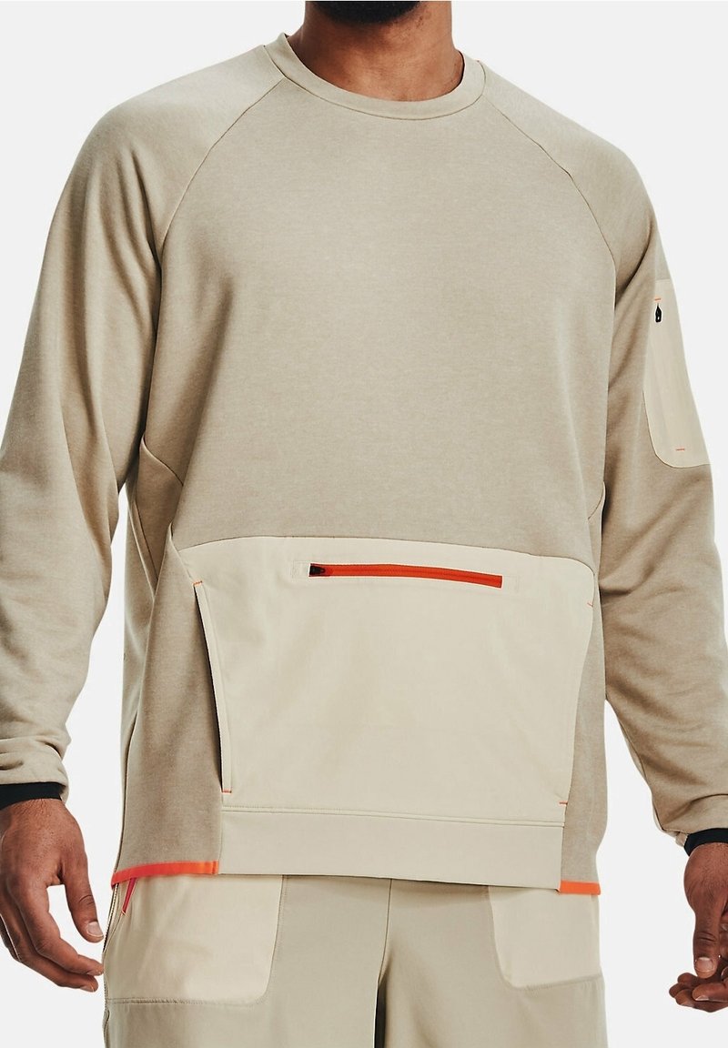 Uomo che indossa un felpa beige con grandi tasche frontali e sulle maniche, entrambe dotate di dettagli arancioni sulle cerniere.