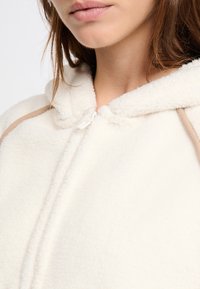 Giacca in pile crema con una texture morbida, caratterizzata da un cappuccio, chiusura a zip e accenti in cordino beige lungo il colletto.