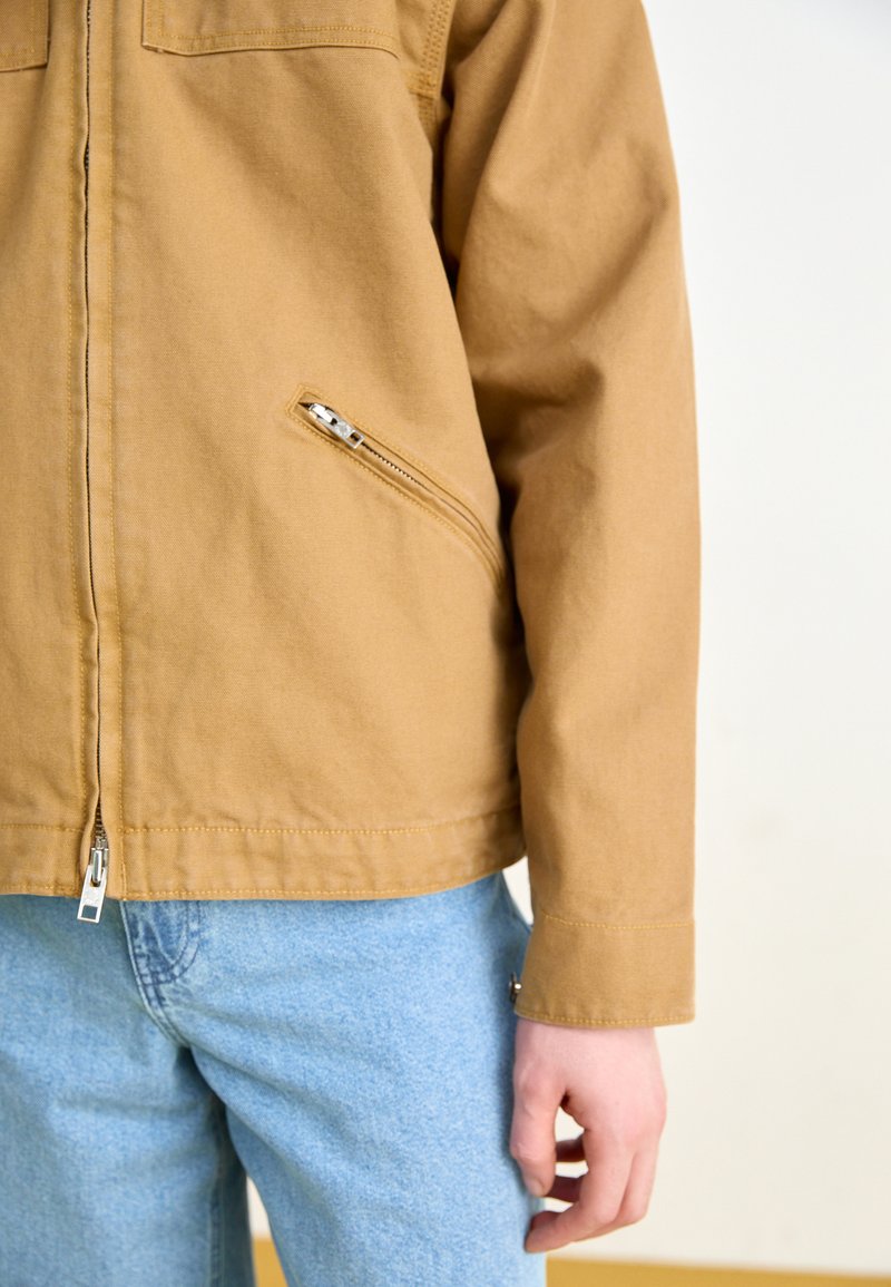 Chaqueta de lona marrón con cremallera frontal y bolsillo lateral en ángulo, combinada con jeans de mezclilla azul claro, mostrando un corte relajado.