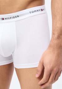 Άσπρα ανδρικά μποξεράκια Tommy Hilfiger, φορεμένα, με ορατή τη ζώνη μέσης με το όνομα και το λογότυπο της μάρκας, καθώς και μερική θέα του δεξιού χεριού και του μηρού.
