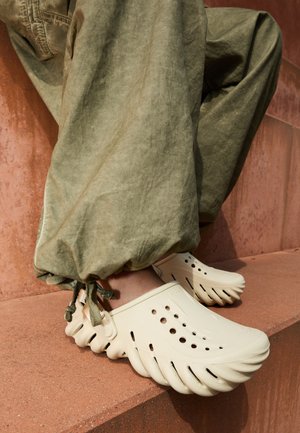 Beige rubber clogs with a wavy edge and ventilation holes, worn with green, cinched cuff pants, resting on a textured terracotta surface.
