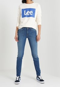 Sudadera crema con logo "Lee" en azul, jeans ajustados azules y zapatillas negras y blancas. Conjunto casual con diseño simple y contraste de colores.