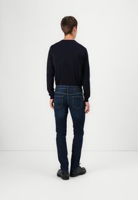 Man som står vänd mot en vit vägg, iklädd en marinblå tröja, mörkblå slim-fit jeans och svarta snörskor.