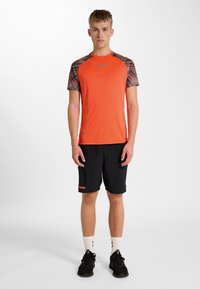 Orange atletisk t-shirt med mönstrade ärmar, svarta shorts, svarta skor och vita strumpor, designad för sport eller vardagsbruk.