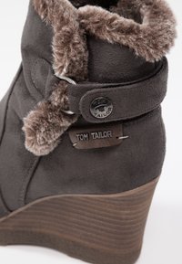 Stivale wedge in suede grigio scuro con bordo in pelliccia sintetica, accenti metallici e una etichetta con logo. Suola wedge testurizzata con strisce orizzontali.
