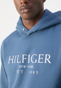 Felpa con cappuccio blu realizzata in tessuto morbido, con un logo bianco che recita "HILFIGER NEW YORK EST. 1985," e un dettaglio con bottone a pressione sul collo.