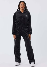 Chándal de velour negro que incluye una chaqueta con capucha y cremallera, con bolsillos frontales, y pantalones a juego con cintura elástica. Tejido texturizado con un acabado suave.