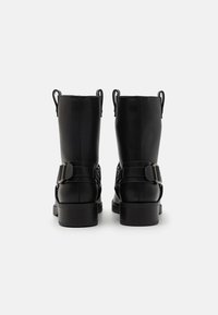 Botines de cuero negro con acabado texturizado, presentan un tacón grueso y correas ajustables con hebillas a los lados. Dos lengüetas en la parte trasera.