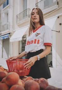 Wit gestreepte jersey met rode accenten en een logo, gecombineerd met een zwarte rok. De persoon houdt een rode mand vol fruit vast.