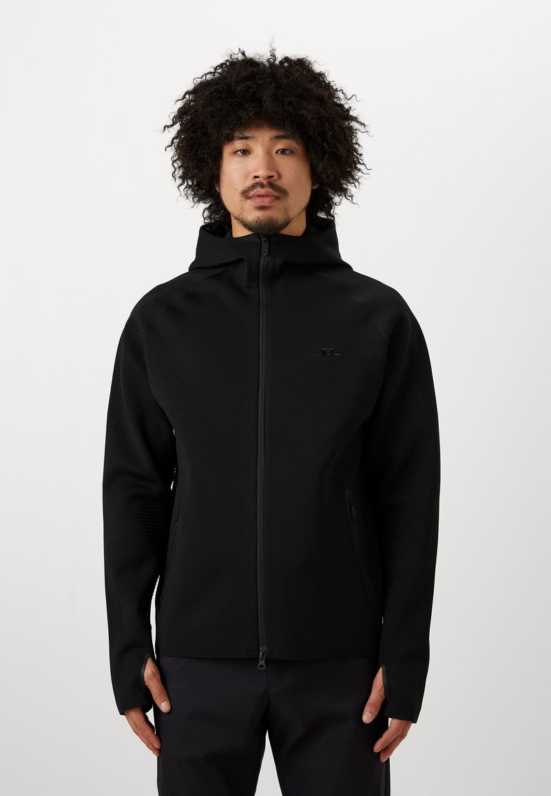 Chaqueta negra con cremallera y capucha, con textura acanalada, bolsillos laterales y un logotipo minimalista en el pecho, llevada sobre pantalones oscuros.