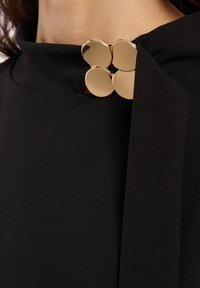 Vêtement en tissu noir avec un col haut, orné d'une accent en or composé de quatre disques circulaires au niveau de la fermeture, ajoutant un détail élégant.