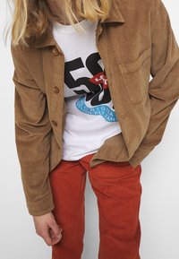 Personne portant une veste en velours côtelé beige, un t-shirt graphique blanc avec le texte "550" et un pantalon en velours côtelé orange brûlé devant un fond uni.