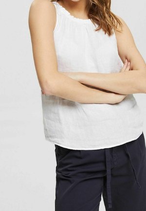 Kobieta w białej, bezrękawnikowej bluzce i ciemno granatowych spodniach, ze skrzyżowanymi ramionami, kadrowana od szyi do talii na jednolitym tle.