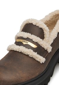 Brauner Slipper mit flauschigem cremefarbenem Innenfutter und goldfarbenem Logo-Detail. Strukturiert Oberfläche und dicke schwarze Sohle für Stabilität.
