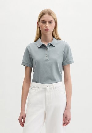 Młoda kobieta ubrana w jasnoniebieską koszulkę polo z krótkim rękawem i białe spodnie z wysokim stanem, stojąca na jednolitym białym tle.