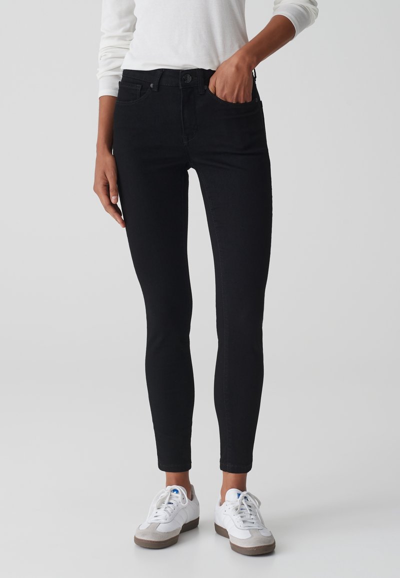 Personne portant un jean noir ajusté, une chemise blanche à manches longues et des baskets blanches avec des détails beige, debout devant un fond uni.