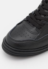 Baskets en cuir noir avec boîte à orteils perforée, accents noirs texturés, lacets plats noirs et semelle en caoutchouc présentant des éléments de design superposés.