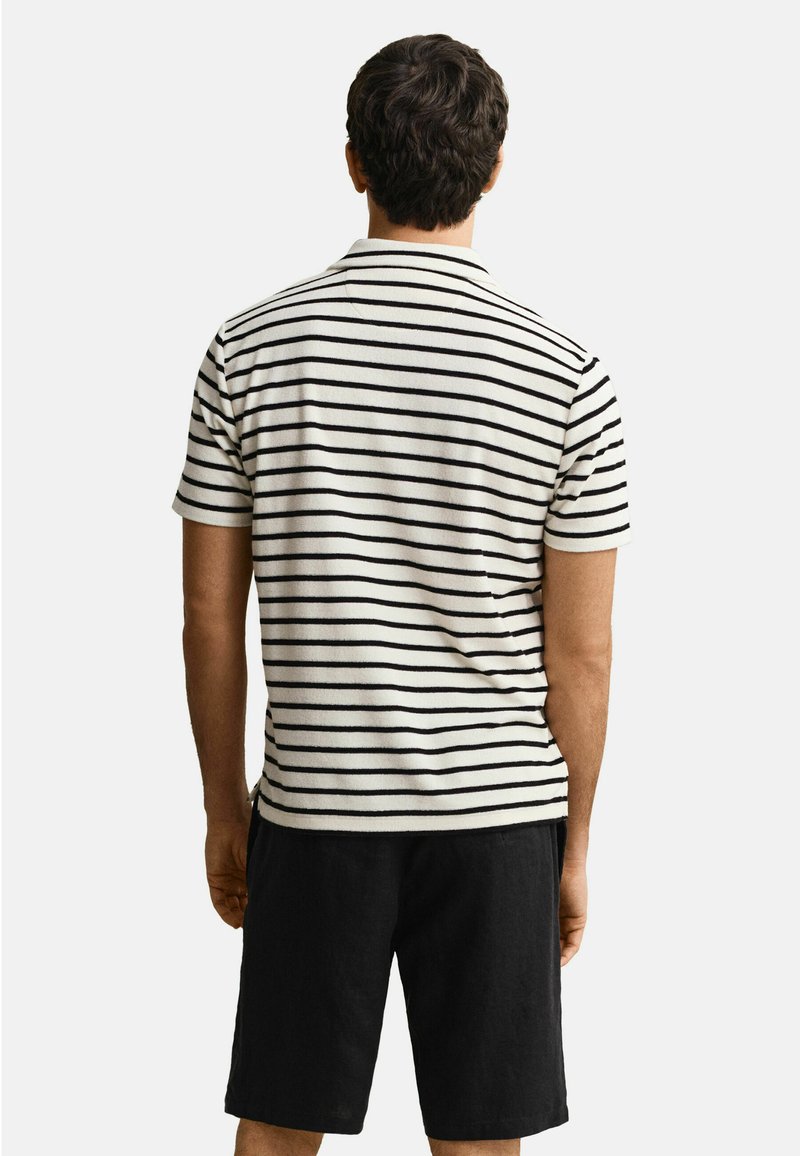 Homme de dos portant une chemise blanche à manches courtes avec des rayures horizontales noires et un short noir arrivant au genou.