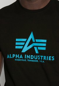 Schwarzes Baumwoll-T-Shirt mit leuchtend blauem "ALPHA INDUSTRIES"-Logo, das ein stilisiertes "A" und horizontale Streifen zeigt. Der Text enthält Standortdetails.