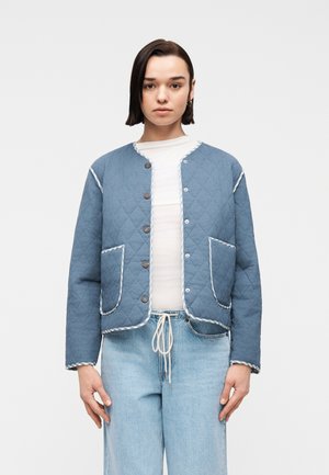 Junge Frau mit kurzen dunklen Haaren, die eine blaue gesteppte Jacke über einem weißen Hemd und hellblauen Jeans trägt und vor einem weißen Hintergrund steht.