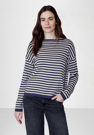Femme aux longs cheveux bruns portant un pull à manches longues rayé bleu marine et blanc et un jean foncé, debout avec une main dans la poche.