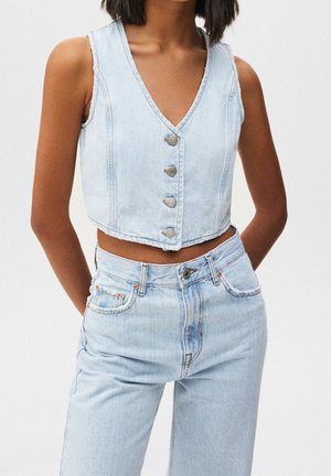 Femme portant un crop top en jean bleu clair sans manches et boutonné, ainsi qu'un jean taille haute, les mains dans le dos.