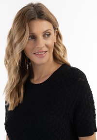 Sort kortærmet sweater med en struktureret bølgedesign, rund halsudskæring og ribbede kanter, sat mod en hvid baggrund.