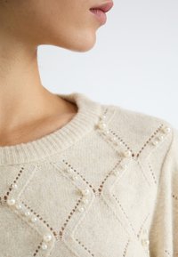 Pull en maille beige avec un col rond côtelé, des motifs en losange et des perles décoratives le long des coutures pour ajouter de la texture.