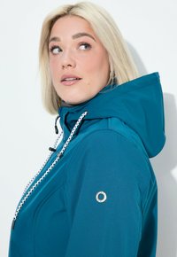 Veste imperméable teal avec un design ajusté, capuche ajustable, cordon de serrage à motifs blancs et noirs, et un accent métallique rond sur la manche.