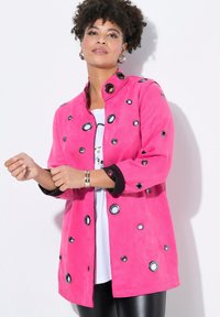 Růžová semišová bunda s kulatými černobílými ozdobnými kroužky, mandarínským límcem a rukávy tříčtvrťového střihu, kombinovaná s bílým topem.