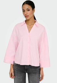 Jeune femme portant des boucles d'oreilles en créole, une chemise surdimensionnée rose clair boutonnée et un jean noir délavé, posant devant un fond uni.