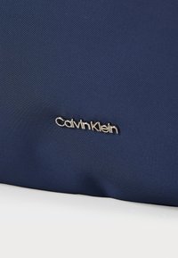 Marineblåt struktureret stof med en tekstureret overflade, prydet med et sølvfarvet Calvin Klein-logoaccents i nederste venstre hjørne.