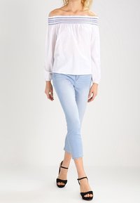 Blouse blanche à épaules dénudées avec smocks bleus, manches longues, associée à un jean court bleu clair et des talons noirs à brides.