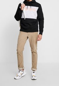 Sudadera negra y gris con logo, pantalones de pana beige y zapatillas gris y blancas con detalles amarillos. Sostiene una mochila ligera.