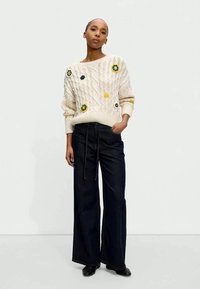 Cream cable-knit sweater with floral embroidery, paired with high-waisted dark denim wide-leg trousers. Black shoes complete the outfit.