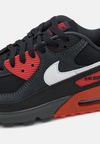 Sneaker nera con accenti rossi e bianchi, sovrapposizioni testurizzate e un'unità di ammortizzazione ad aria visibile. Presenti lacci e dettagli del marchio.