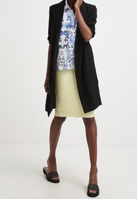 Un lungo cappotto nero su una camicia floreale blu e bianca, abbinato a una gonna rigata giallo chiaro e sandali neri con suola piatta.